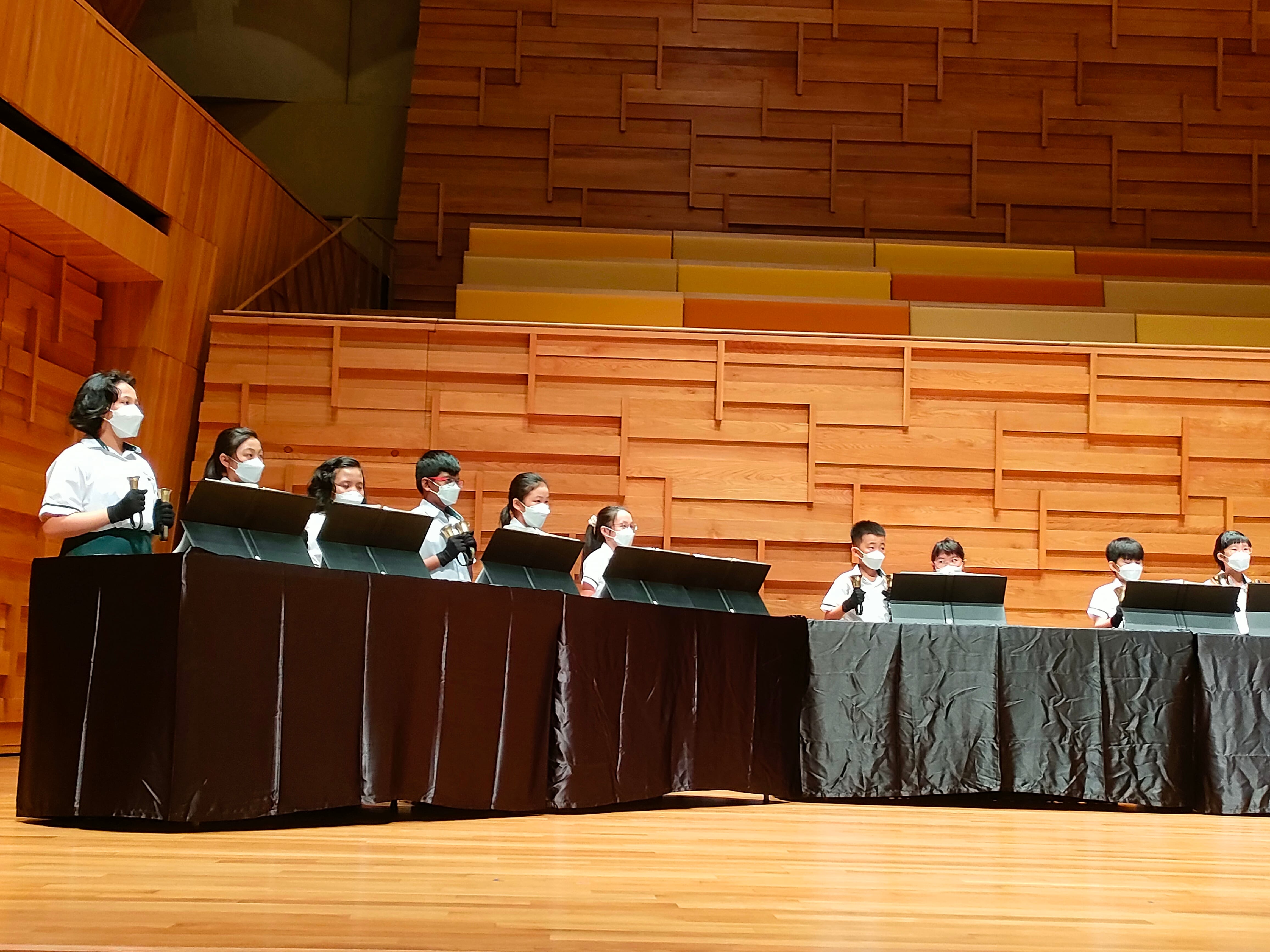 Children wearing white uniforms and face masks stand at draped tables playing handbells on a wooden concert stage.